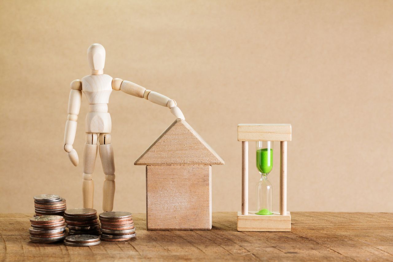 Wooden mannequin with house model, coins, and hourglass symbolizing financial planning.